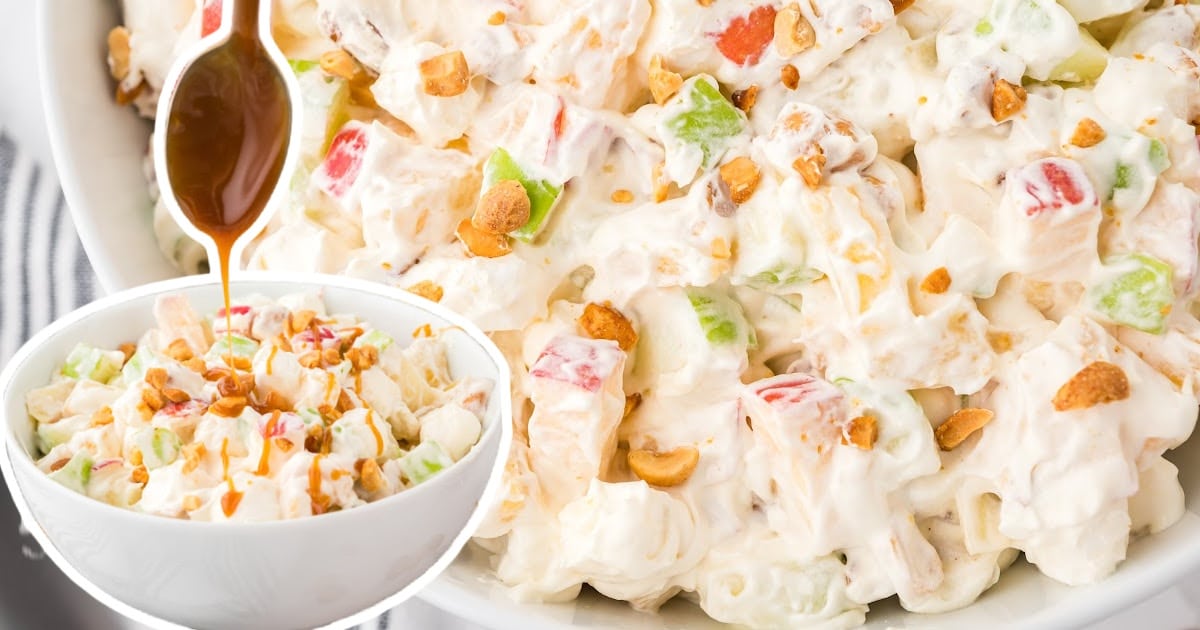 A bowl of creamy taffy apple salad filled with diced red and green apples, mini marshmallows, peanuts, and vanilla pudding, being drizzled with rich caramel sauce from a spoon overhead. Other bowls of salad and scattered peanuts are visible in the background.