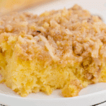 A close-up of a moist, fluffy slice of Tornado Cake topped with a golden coconut and pecan mixture, served on a small white plate with more cake visible in the background.