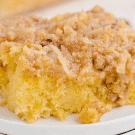 A close-up of a moist, fluffy slice of Tornado Cake topped with a golden coconut and pecan mixture, served on a small white plate with more cake visible in the background.