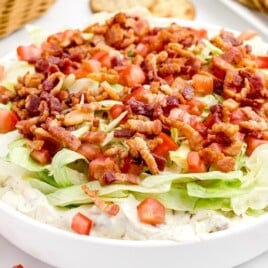 Close-up of creamy BLT dip topped with chopped bacon, fresh tomatoes, and crisp lettuce, served in a white bowl with crackers on the side.