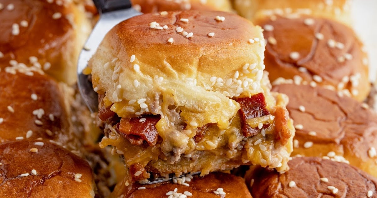 Baked bacon cheeseburger sliders with melted cheddar cheese and bacon layered between soft sesame slider buns, fresh from the oven in a glass baking dish.