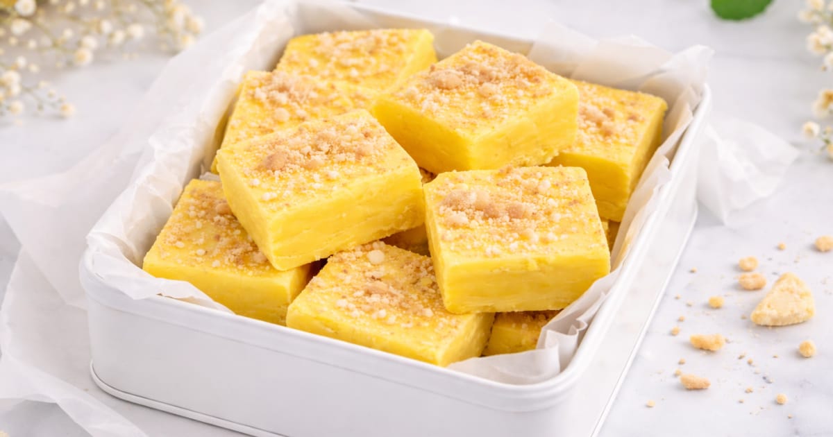 Banana fudge squares arranged in a white rectangular gift tin lined with parchment paper, topped with crushed vanilla wafer crumbs, set on a light marble surface with soft decorative greenery in the background
