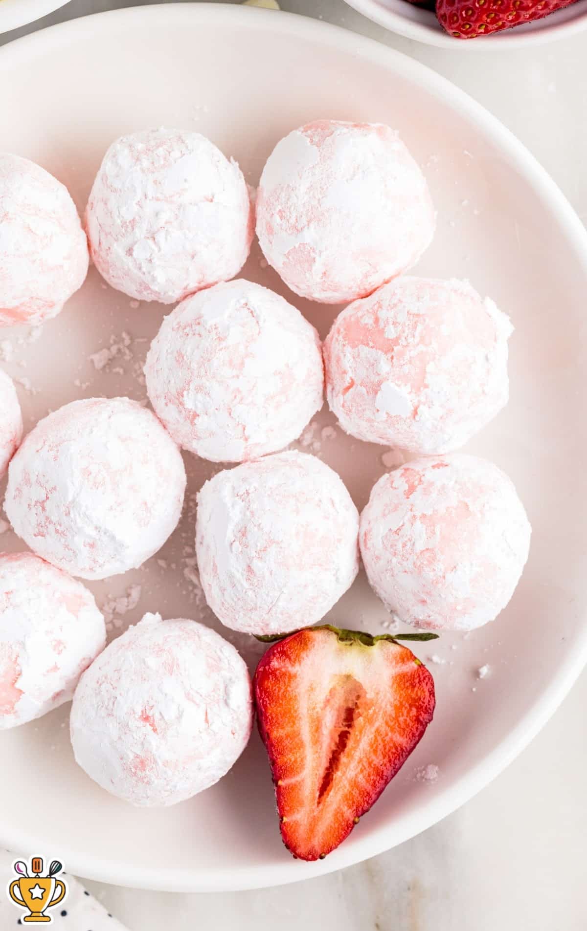 Strawberry truffles stacked high on a white plate, lightly dusted with powdered sugar, with a bowl of fresh strawberries nearby for garnish.