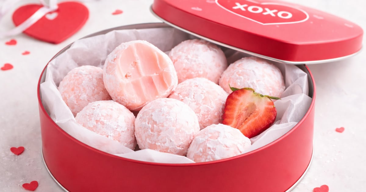 Pink strawberry truffles dusted with powdered sugar arranged inside a deep red Valentine’s Day treat tin lined with parchmented parchment paper, with one truffle cut open to show the creamy strawberry center and a fresh sliced strawberry tucked alongside for garnish.