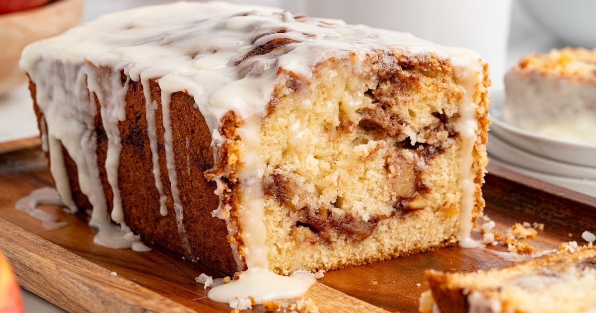 A glazed apple fritter bread loaf with one thick slice cut out, showing moist yellow cake layered with cinnamon-spiced apples, sitting on a wooden board with white glaze dripping over the top and pooling slightly, styled in a bright kitchen setting.