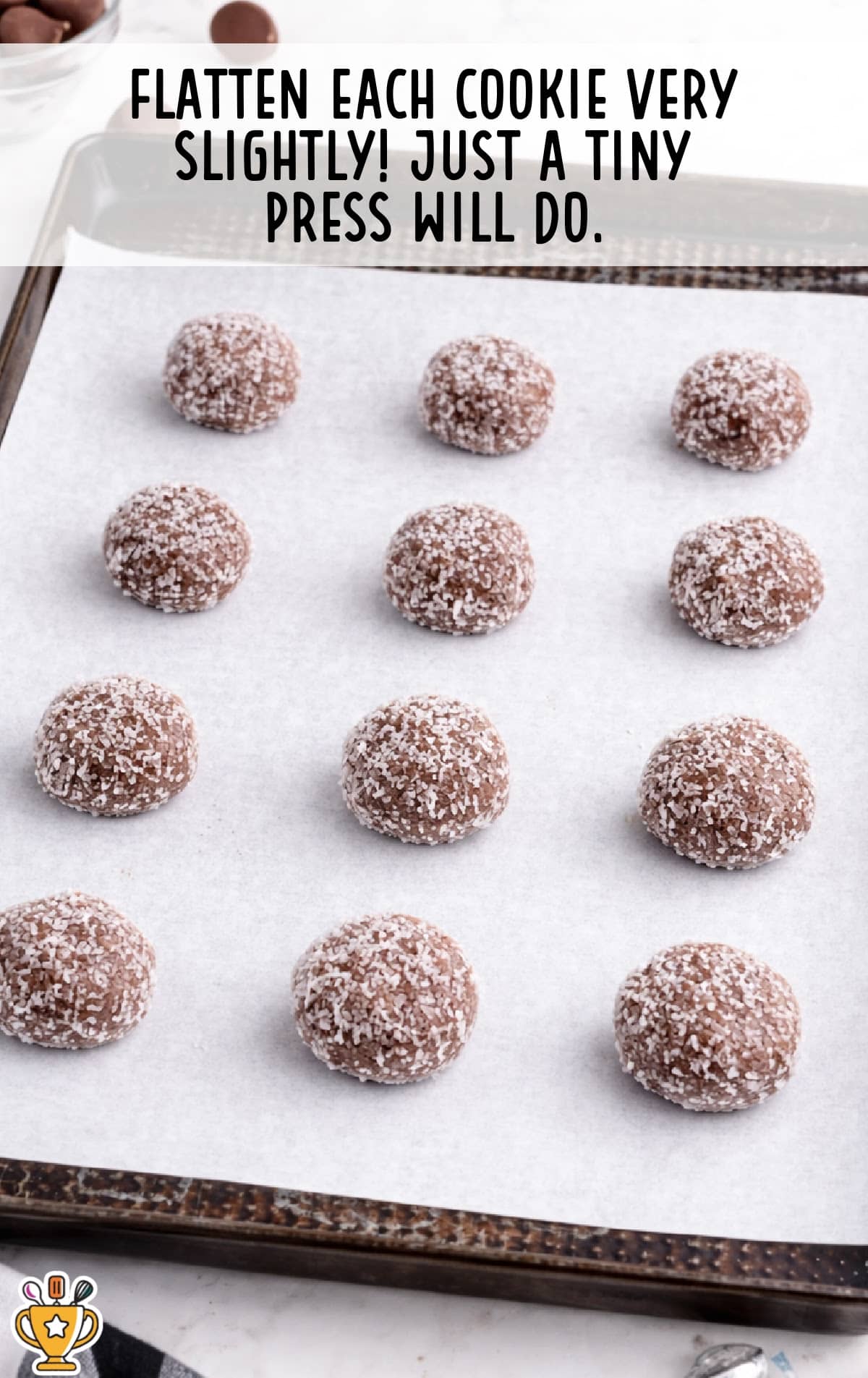 Chocolate sugar cookie dough balls rolled in coarse sanding sugar and placed on a parchment-lined baking sheet, with text reading “Flatten each cookie very slightly, just a tiny press will do.”