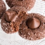 Chocolate sugar kiss cookies on a glass plate, one cookie broken open to show a soft, puffy chocolate interior with a Hershey’s Kiss pressed into the center.