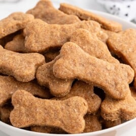 A close-up feature image of stacked, bone-shaped homemade dog treats inside and spilling out of a clear glass jar with a black label that reads “DOG TREATS.” The treats are golden brown with a slightly rough, whole-grain texture. The background is bright and neutral with a clean kitchen aesthetic.