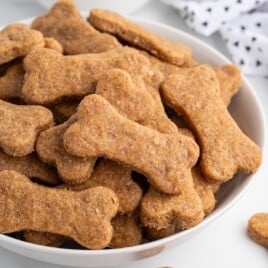 A close-up recipe card image featuring a white bowl filled with baked, bone-shaped homemade dog treats stacked and overlapping. The treats are golden brown with a visible whole-grain texture and slightly rounded edges. The background is bright and clean, resembling a light kitchen surface with a folded white cloth patterned with small black shapes partially visible in the background.
