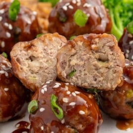 Close-up of glazed Asian turkey meatballs topped with sesame seeds and green onions, with one meatball cut open to show the juicy inside, served with rice and broccoli.