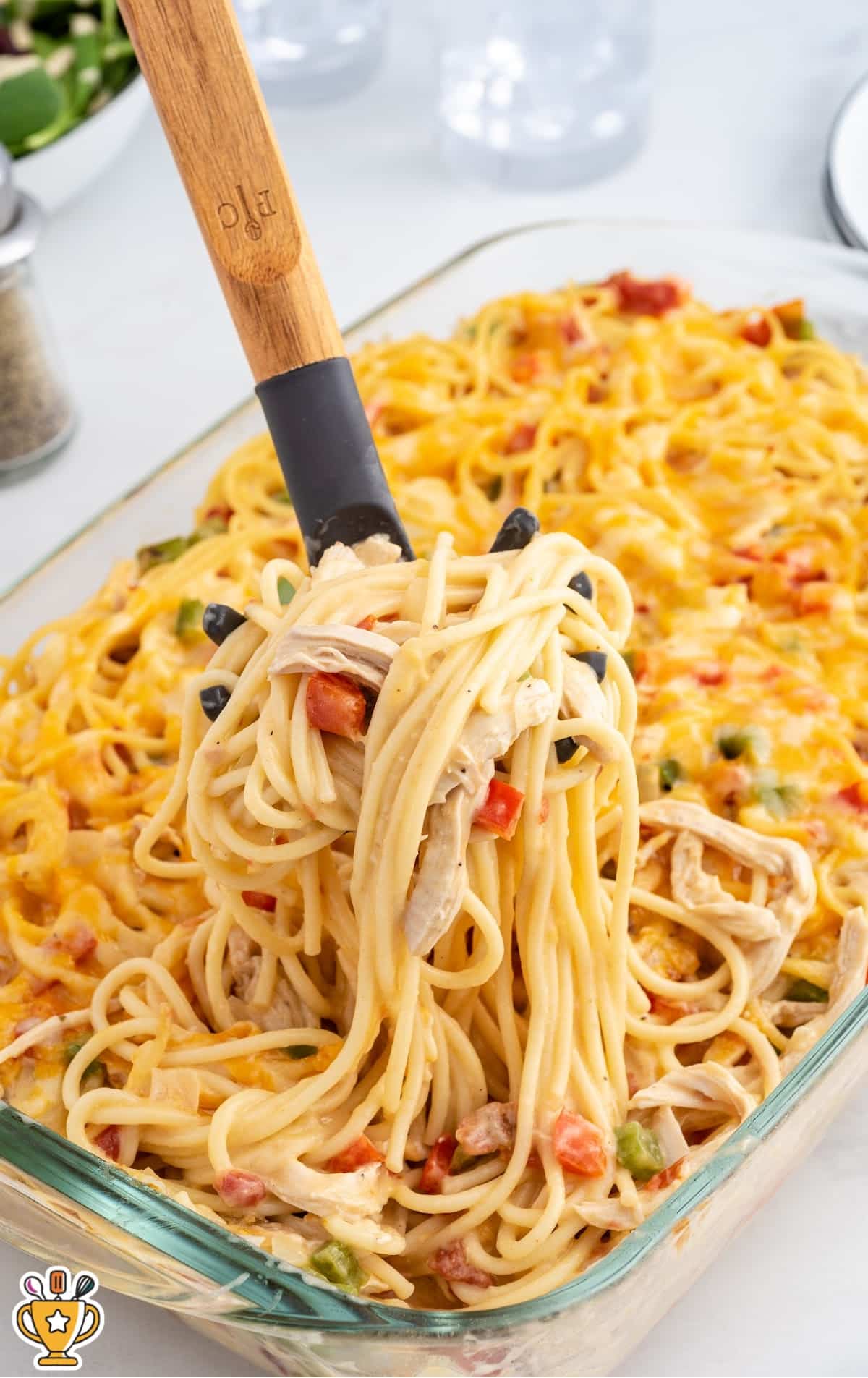 Close-up of cheesy baked chicken spaghetti with shredded chicken and diced peppers being lifted from a glass baking dish with a pasta server.