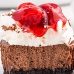 Close-up of a Black Forest mini cheesecake with a chocolate cookie crust, chocolate cheesecake layer, whipped topping, and glossy cherry pie filling dripping down the center. Chocolate shavings are sprinkled on top. Text on the image reads “The Yummiest Black Forest Mini Cheesecakes” and “Topped with Juicy Cherries,” with a can of cherry pie filling shown beside the cheesecake.