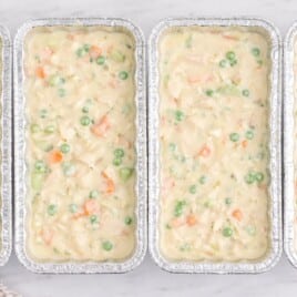 Four unbaked chicken pot pies filled with creamy chicken, peas, carrots, and celery arranged side by side in foil loaf pans on a white marble surface.