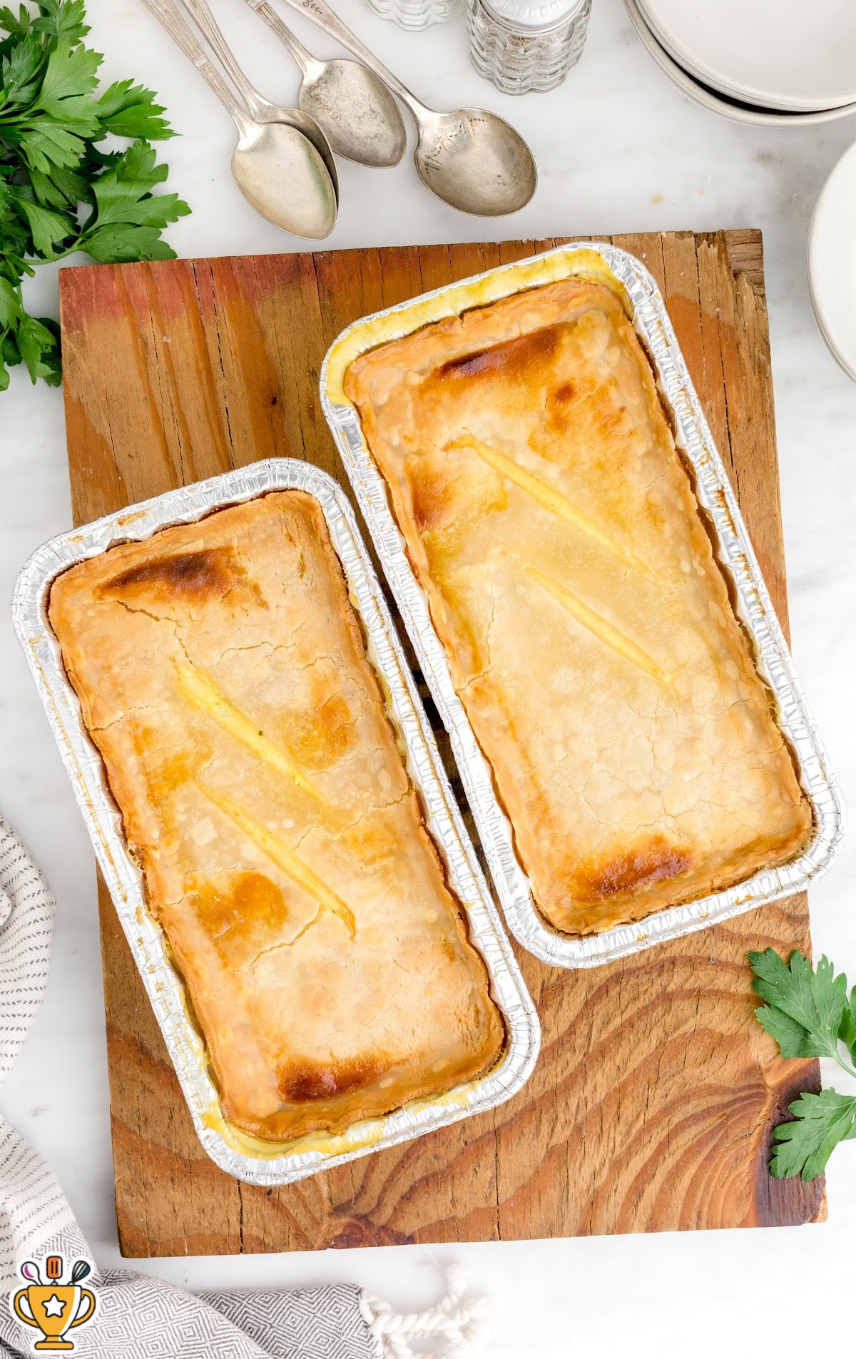 Overhead view of two baked freezer chicken pot pies in disposable foil loaf pans with golden, flaky crusts, set on a wooden cutting board. Fresh parsley and silverware are styled around the board, highlighting the finished make-ahead chicken pot pies.