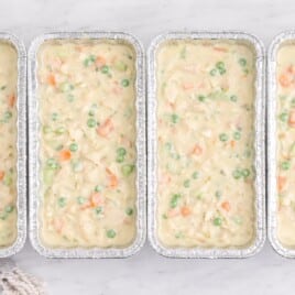 Row of unbaked freezer chicken pot pies filled with creamy chicken and vegetables in foil loaf pans on a white marble surface.