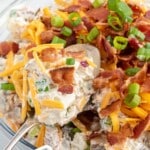Overhead view of loaded baked potato salad in a glass bowl, finished with bacon, cheese, and green onions, styled as a vertical pin image.