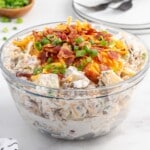 Finished loaded baked potato salad in a clear bowl, fully mixed with creamy dressing and topped with bacon, shredded cheddar cheese, and green onions.