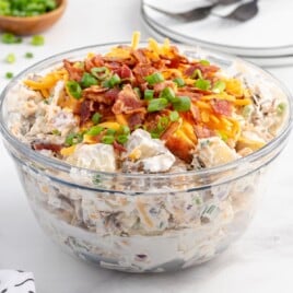 Finished loaded baked potato salad in a clear bowl, fully mixed with creamy dressing and topped with bacon, shredded cheddar cheese, and green onions.