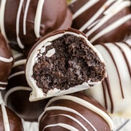 Close-up of bite-sized Oreo Balls coated in chocolate with white chocolate drizzle, stacked together on a white serving plate. One Oreo ball is cut open in the center, clearly showing the rich Oreo cookie filling inside. Text on the image reads “Bite-Sized Oreo Balls” and “Made with only 5 I
