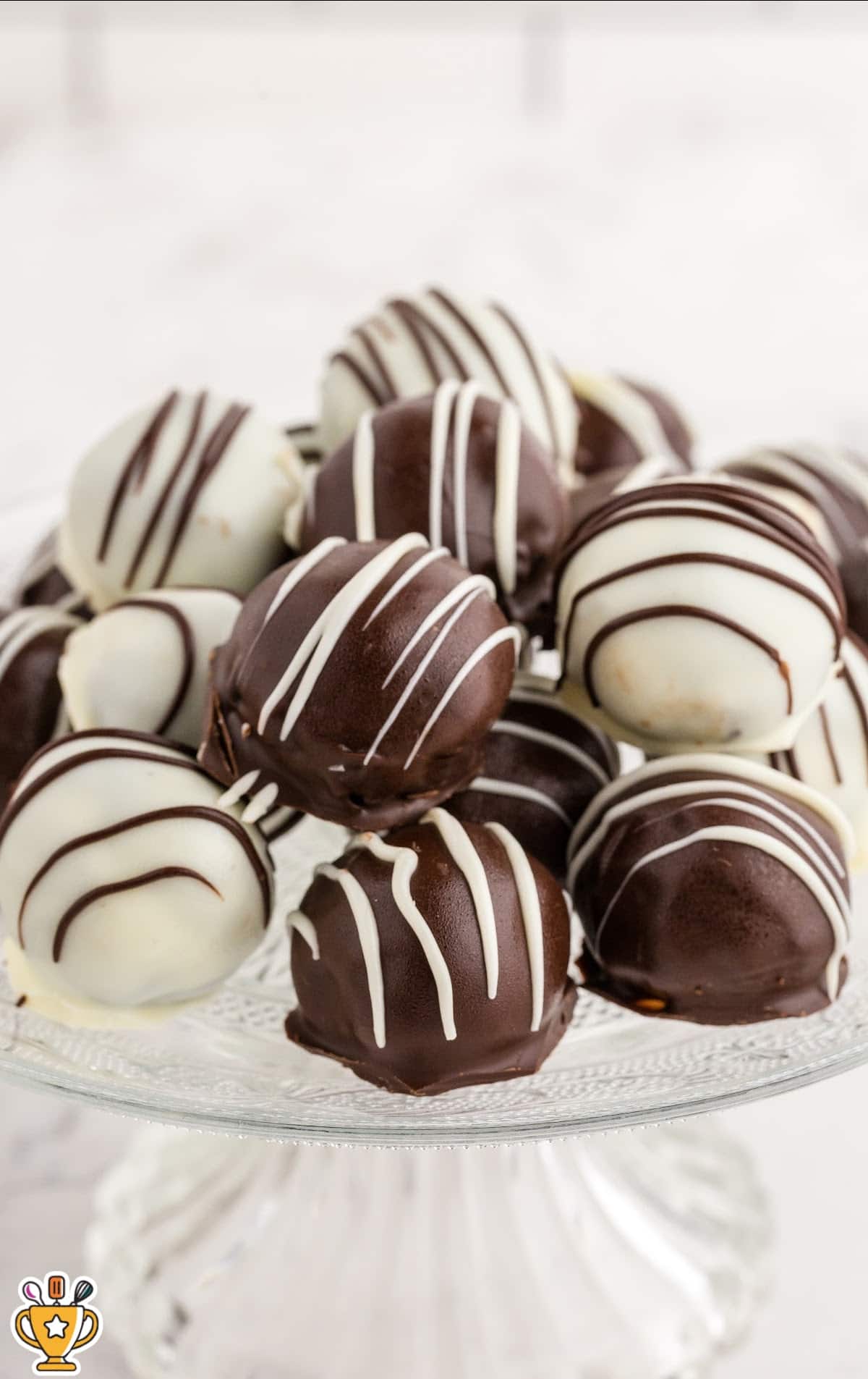 Chocolate and white chocolate Oreo Balls displayed on a white pedestal cake stand with a clean, light background. The Oreo Balls are evenly drizzled and arranged in a neat pile, showcasing their glossy chocolate coating and bite-sized shape.