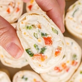 Hand holding a ranch roll up pinwheel slice over a tray of sliced tortilla pinwheels, highlighting the creamy ranch filling with diced red pepper and green onion.