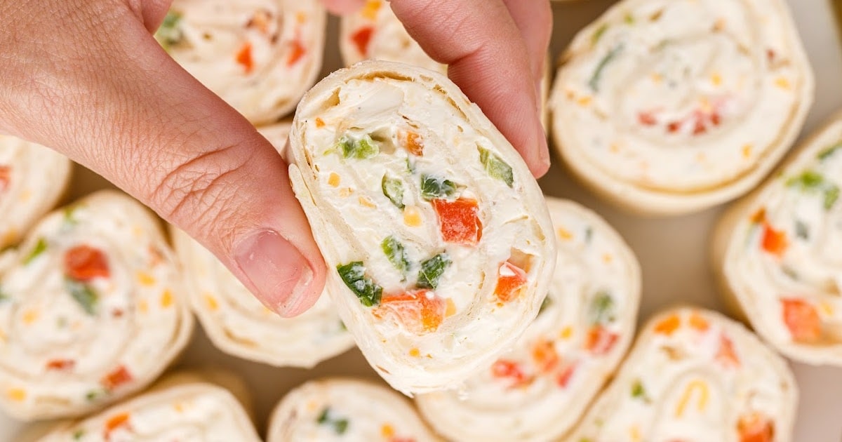 Hand holding a ranch roll up pinwheel slice over a tray of sliced tortilla pinwheels, highlighting the creamy ranch filling with diced red pepper and green onion.