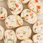 Overhead close-up of sliced Ranch Roll Ups arranged on a neutral surface, showing soft flour tortilla pinwheels filled with creamy ranch-seasoned cream cheese, diced red or orange bell peppers, green onions, jalapeño, and bits of cheddar jack cheese.