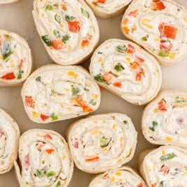 Overhead close-up of sliced Ranch Roll Ups arranged on a neutral surface, showing soft flour tortilla pinwheels filled with creamy ranch-seasoned cream cheese, diced red or orange bell peppers, green onions, jalapeño, and bits of cheddar jack cheese.