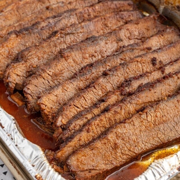 Close-up of slow-cooked brisket sliced and resting in its juices inside a foil-lined roasting pan. The brisket appears moist and tender with visible seasoning and rich cooking liquid pooling around the slices.