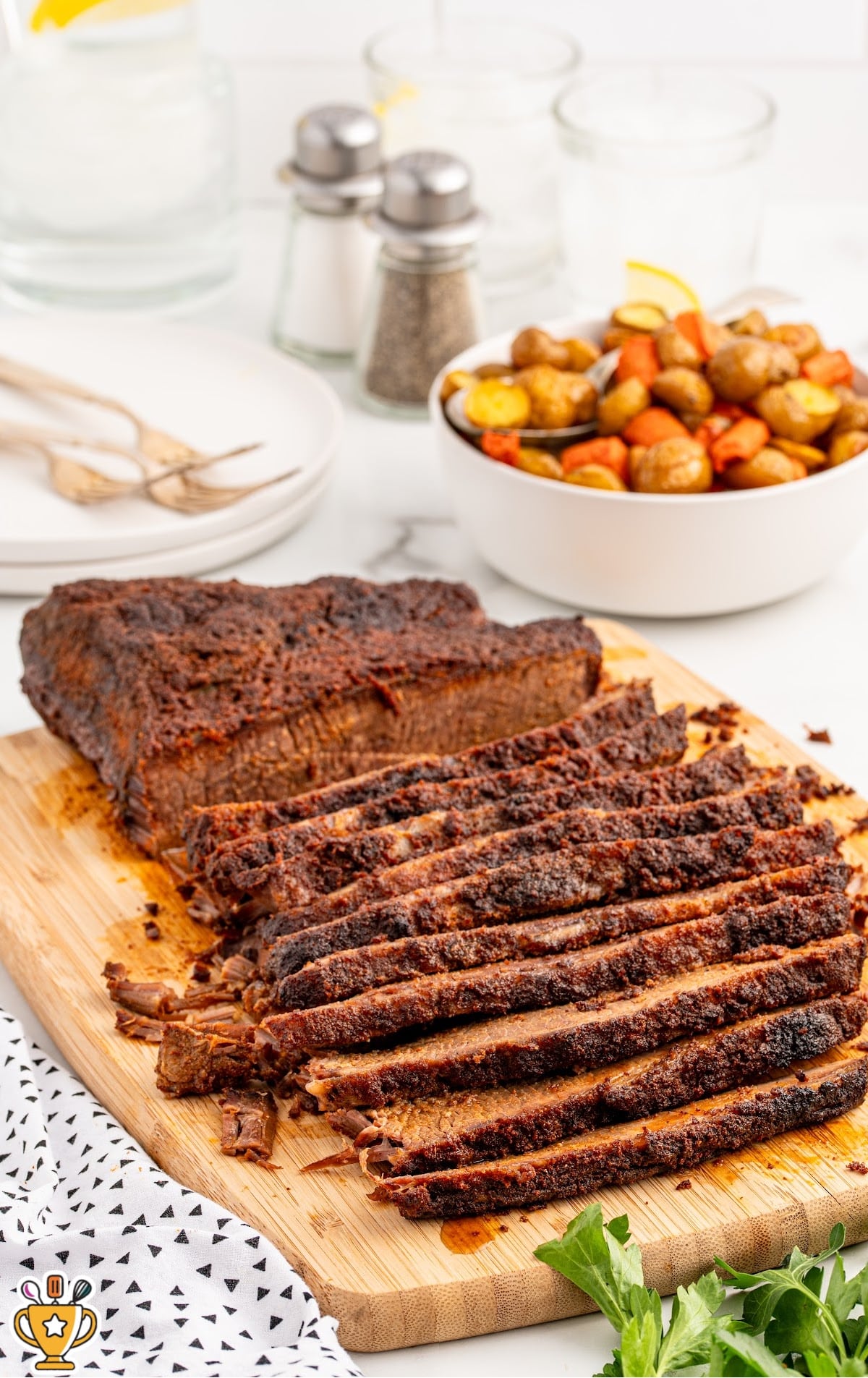 Whole cooked brisket sliced on a wooden cutting board with a dark, flavorful crust. The slices are fanned out to show the juicy interior. In the background are roasted vegetables, stacked plates, salt and pepper shakers, and serving forks, suggesting a complete meal setup.