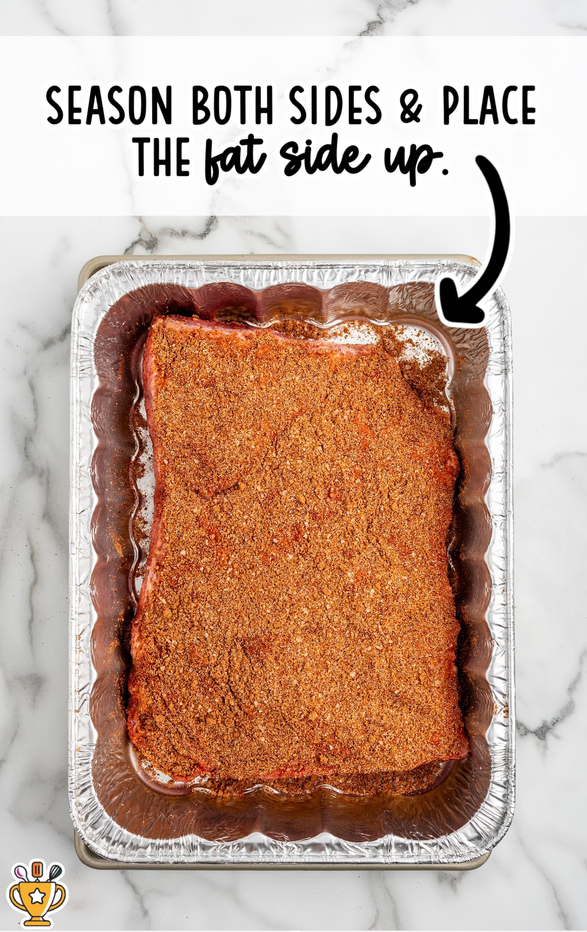 Overhead view of a seasoned beef brisket in a foil pan with the fat side up, showing the rub coating the surface before slow cooking in the oven.