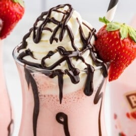 Strawberry Mudslide drink topped with whipped cream and chocolate drizzle, served in a hurricane glass lined with chocolate syrup and garnished with a fresh strawberry and striped straw, with Baileys Strawberries & Cream in the background.