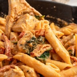 Close-up of creamy tuscan chicken pasta in a black skillet, with penne, tender chicken pieces, spinach, and sun-dried tomatoes, with a wooden spoon lifting the pasta.