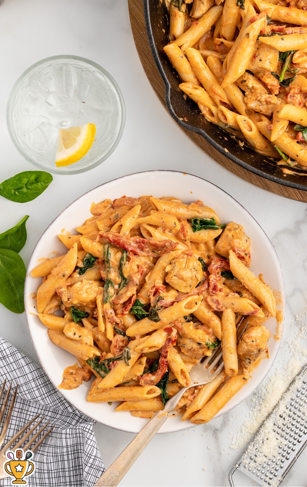 Plated serving of tuscan chicken pasta with penne, chicken, spinach, and sun-dried tomatoes in a creamy sauce, photographed from above with a fork, skillet of pasta in the background, lemon wedge, spinach leaves, and grated cheese on a light surface.