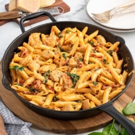 Cast iron skillet filled with creamy tuscan chicken pasta on a wooden board, showing penne, chicken, spinach, and sun-dried tomatoes in a smooth sauce, styled with parmesan cheese, grater, plates, and forks in the background.