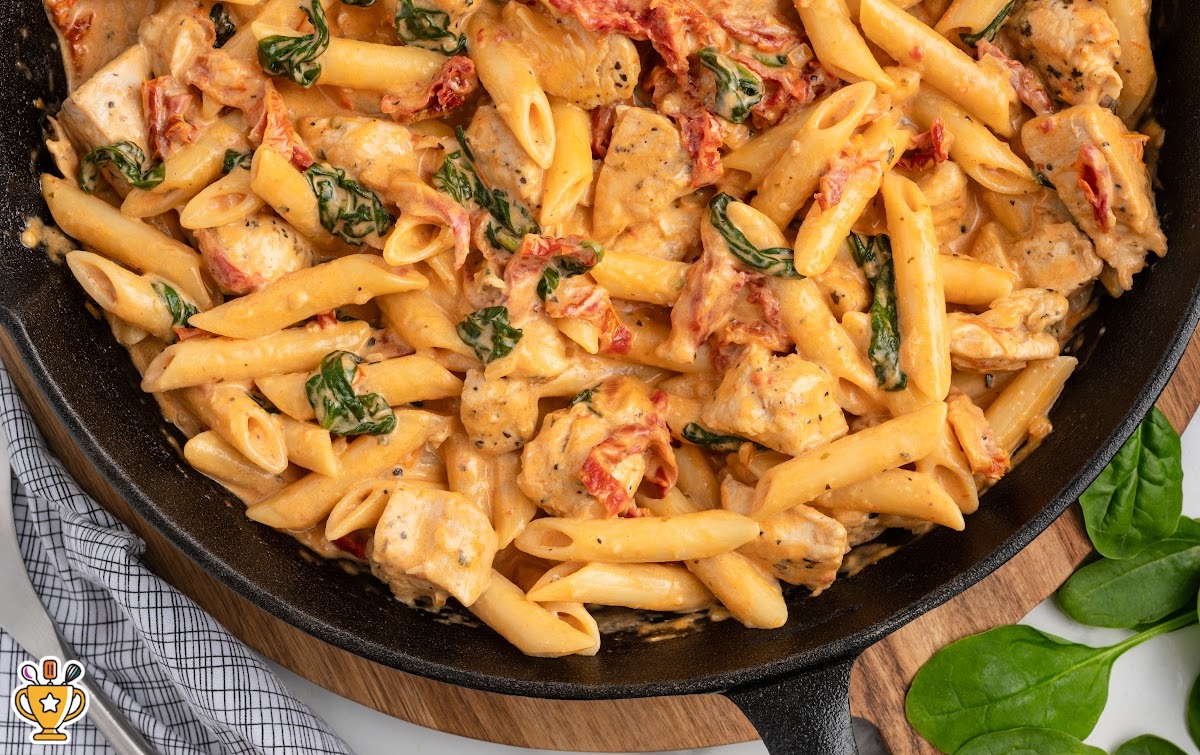 Overhead view of Tuscan chicken pasta in a skillet, showing penne noodles, tender chicken pieces, sun-dried tomatoes, and spinach evenly coated in a creamy parmesan sauce.