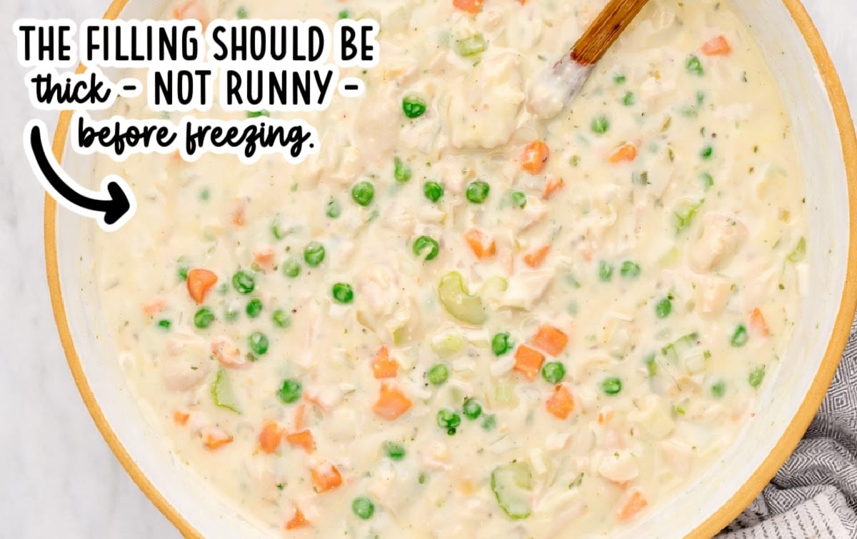 Creamy chicken pot pie filling with chicken, peas, carrots, and celery in a large pan, showing a thick texture, with on-image text indicating the filling should be thick and not runny before freezing.