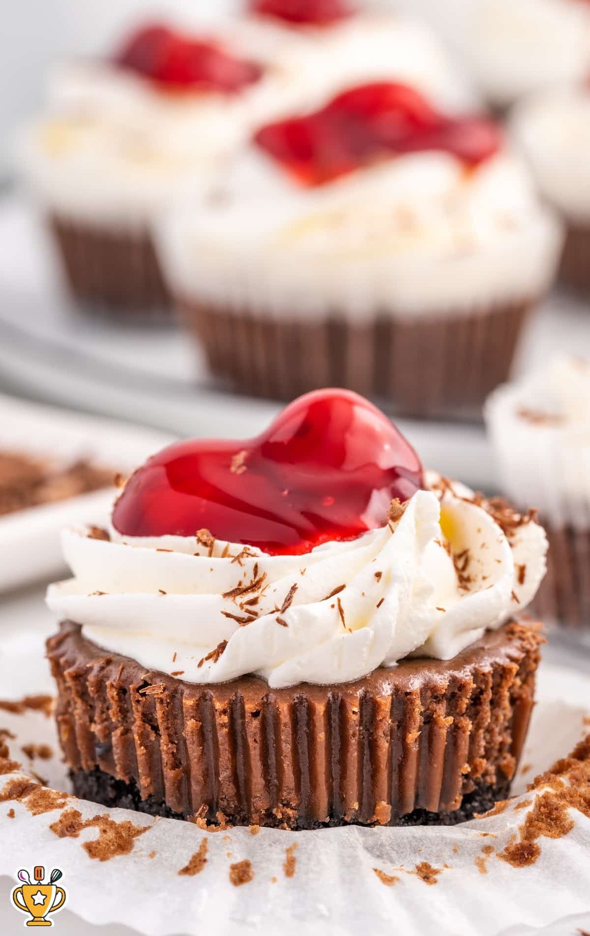 Single black forest mini cheesecake topped with whipped cream, cherry pie filling, and chocolate shavings, sitting on parchment paper with additional mini cheesecakes blurred in the background.