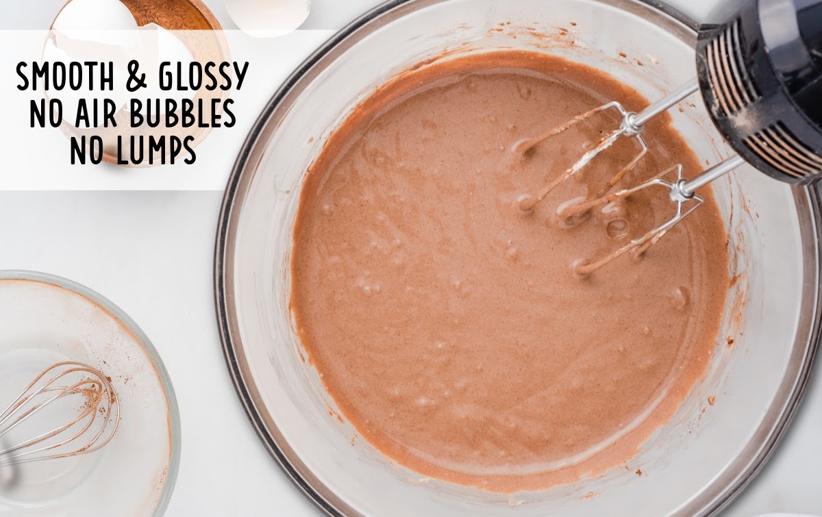 Overhead view of smooth, glossy chocolate cheesecake batter in a mixing bowl with beaters resting inside, showing the correct texture with no lumps or air bubbles, and text reading “Smooth & glossy, no air bubbles, no lumps.”