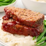 Close-up of two thick slices of Cracker Barrel–style meatloaf stacked on top of each other, coated with a glossy red ketchup glaze. The meatloaf sits on a bed of creamy mashed potatoes with bright green beans on the side. The interior shows a fine, tender crumb with visible bits of green bell pepper and onion.