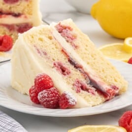 Slice of Lemon Raspberry Cake on a white plate with creamy lemon frosting, layers of raspberry filling and whole raspberries, dusted with powdered sugar. Fresh lemons and raspberries are styled in the background, highlighting the moist texture and vibrant lemon raspberry layers.
