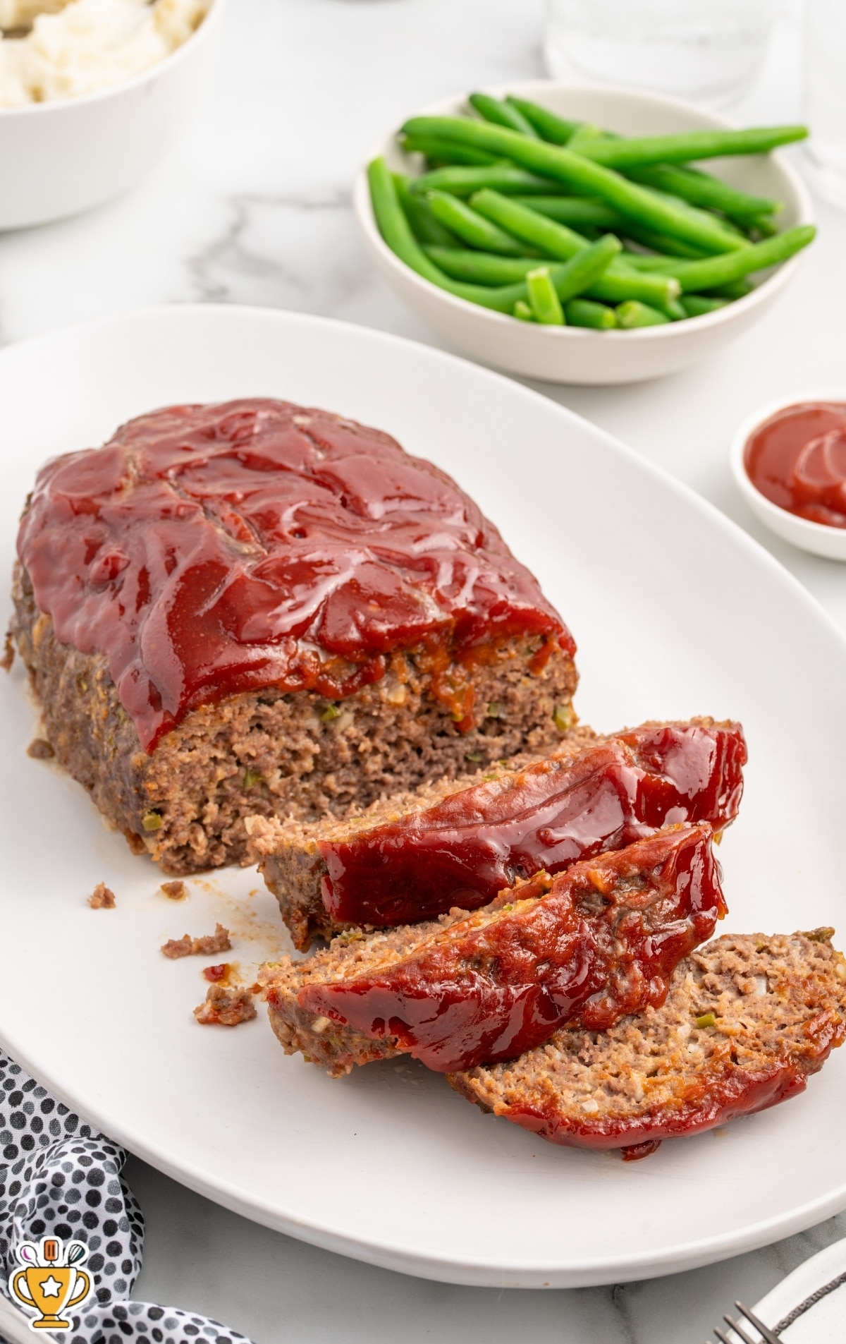 Hero image of a full Cracker Barrel–style meatloaf with one slice cut and placed in front, showing a tender interior dotted with onion and green bell pepper. The loaf is generously coated with a thick red glaze. White bowls of green beans and mashed potatoes sit softly out of focus in the background on a light marble surface.