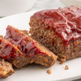 Feature image showing a fork cutting into a slice of Cracker Barrel–style meatloaf covered in a glossy red glaze, served over mashed potatoes with green beans on the side. Text over the image reads “Cracker Barrel Meatloaf” and “with the original savory glaze,” emphasizing the copycat restaurant-style presentation.