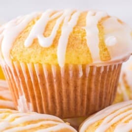 Cake Mix Muffins with smooth white icing drizzle arranged on a plate, featuring a tall lemon muffin in the center and additional muffins in the background, highlighting their tender texture and bakery-style look.