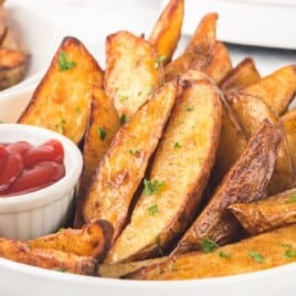 Air fryer potato wedges piled in a white serving bowl with a small ramekin of ketchup, sprinkled with chopped parsley on a bright marble countertop.