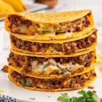 Square food photo of a tall stack of baked tacos layered with seasoned ground beef, melted cheese, and cilantro on a white plate, with cilantro and a patterned kitchen towel near the bottom edge.