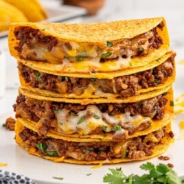 Square food photo of a tall stack of baked tacos layered with seasoned ground beef, melted cheese, and cilantro on a white plate, with cilantro and a patterned kitchen towel near the bottom edge.