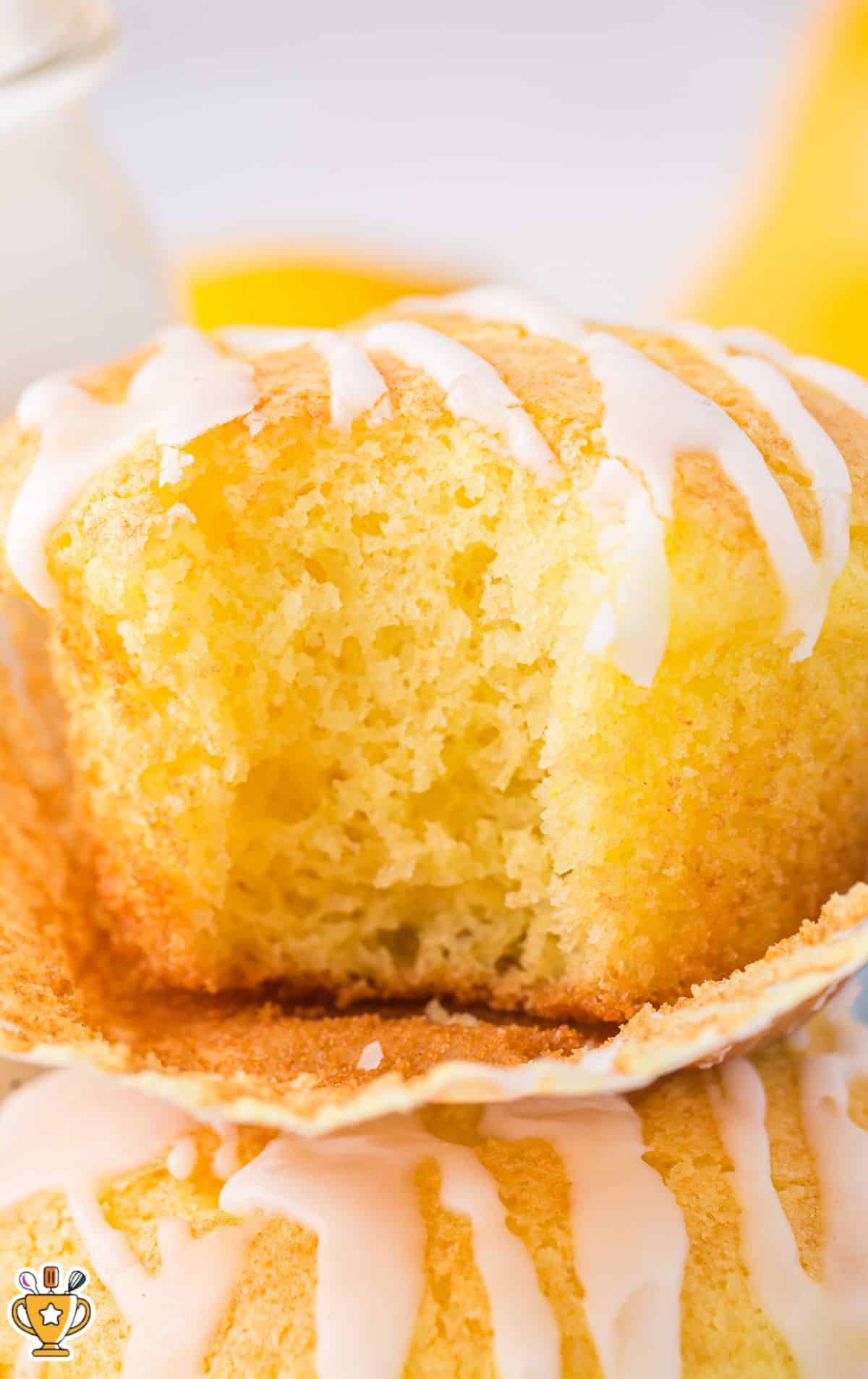 Split lemon Cake Mix Muffin with white glaze drizzle, showing the moist interior crumb and soft texture, stacked with additional glazed muffins in the background for a bright, family-friendly dessert presentation.