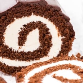 Close-up of a sliced chocolate roll cake showing a thick vanilla cream spiral filling, with another slice in the foreground on a bright white surface.