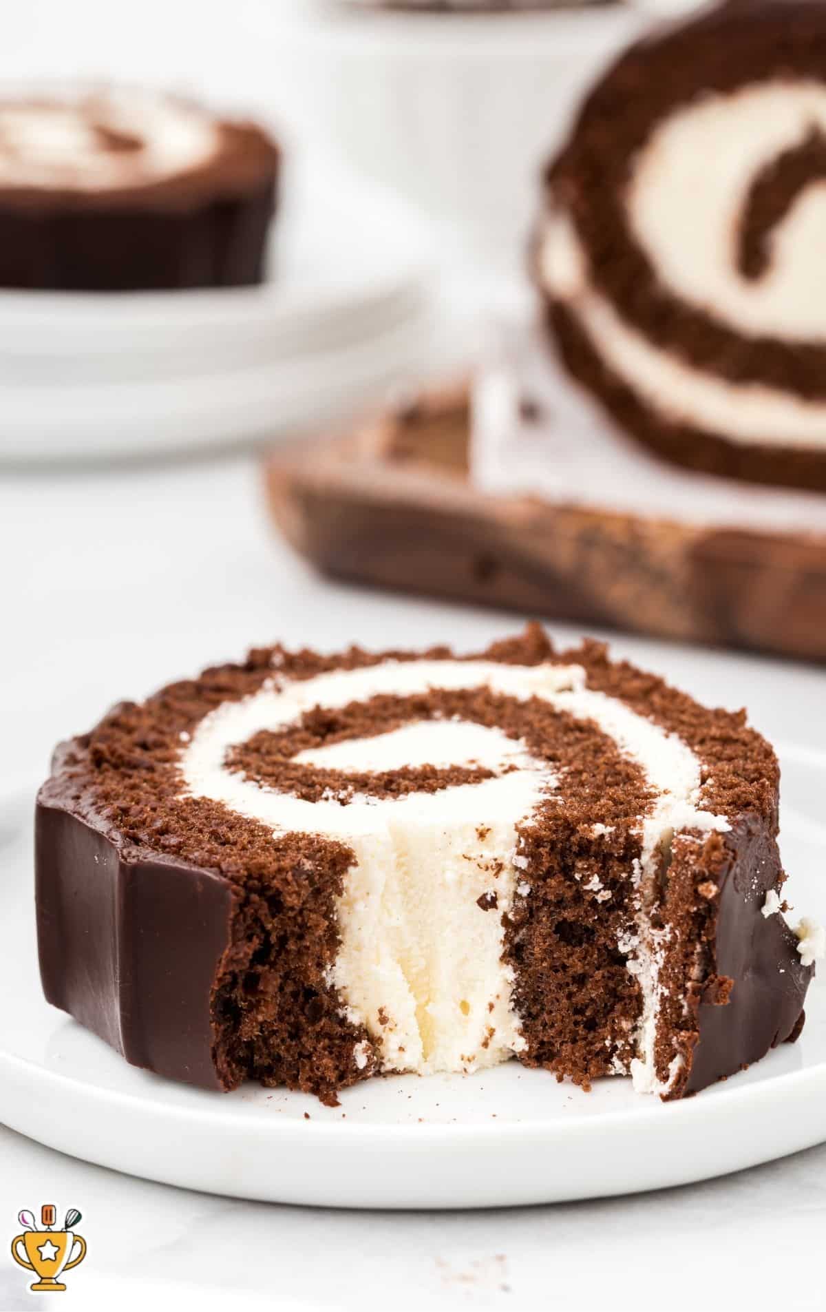 Single slice of chocolate roll cake on a white plate with a bite taken out, showing the vanilla cream spiral and chocolate coating, with more cake blurred in the background.